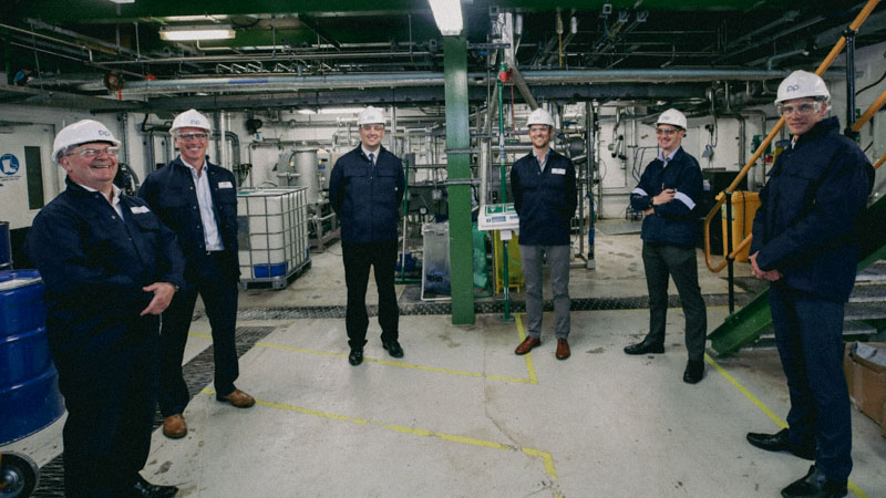A photograph of six men in an industrial environment wearing hard hats and protective overalls