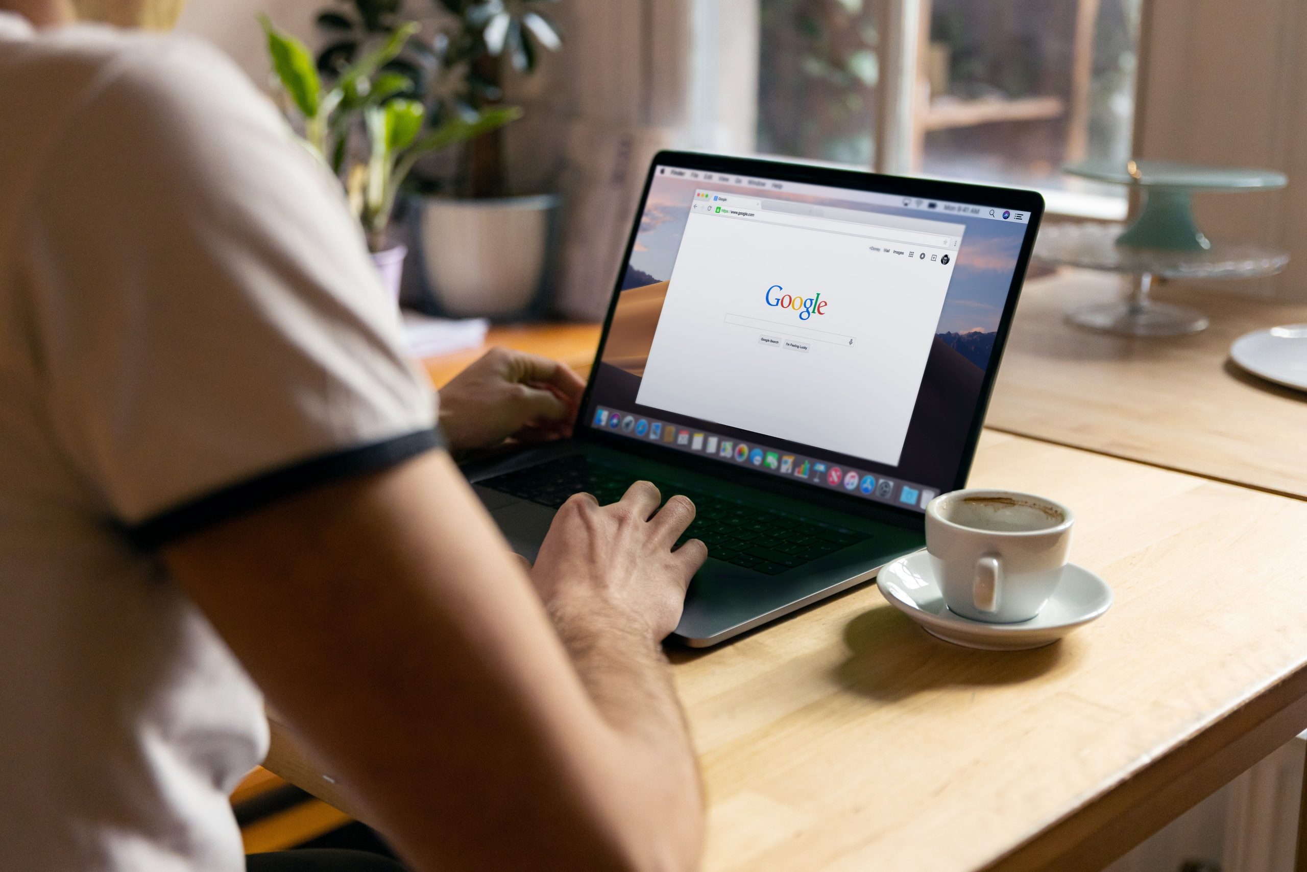 A photograph of a man at a desk using Google on a MacBook Pro with a coffee cup next to him