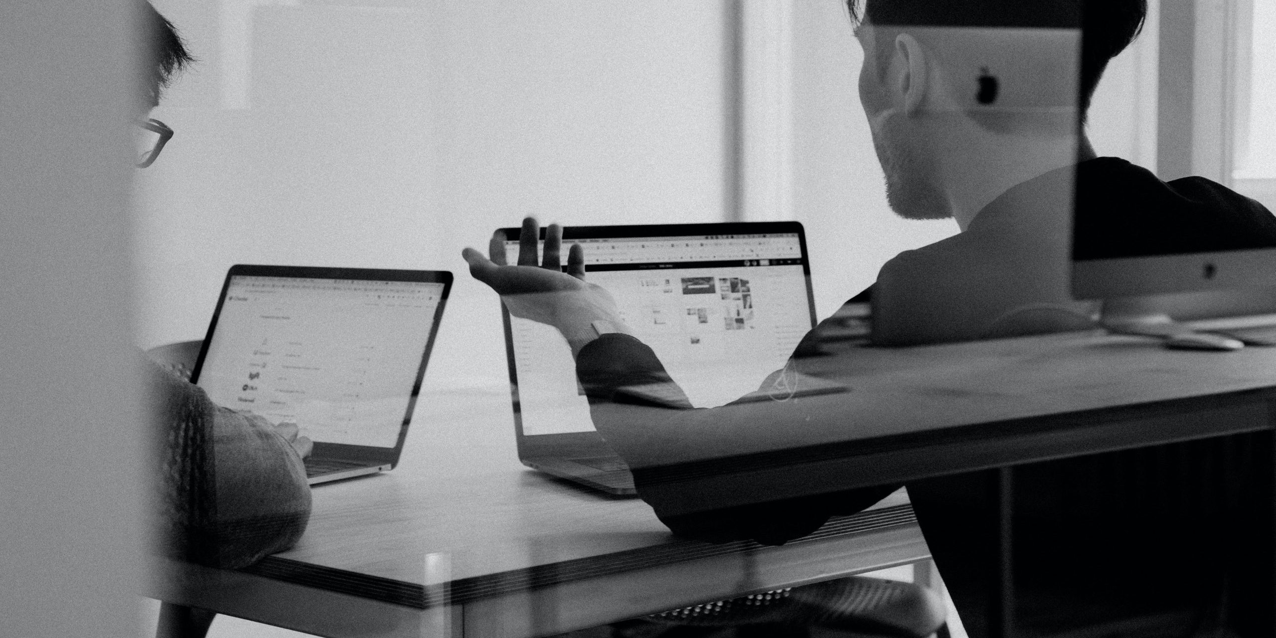 A black and white photograph showing two men in an office on laptops discussing business